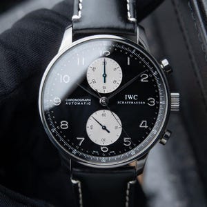 May include: An IWC Schaffhausen automatic chronograph watch with a black dial, white sub-dials, and Arabic numerals. The timepiece features a polished silver-tone stainless steel case and a black leather strap with white contrast stitching.