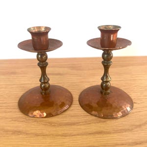 Peut inclure: Deux bougeoirs en cuivre avec une texture martelée. Chaque bougeoir a une base ronde, une tige décorative et une petite coupelle pour une bougie. Les bougeoirs sont présentés sur une surface en bois.