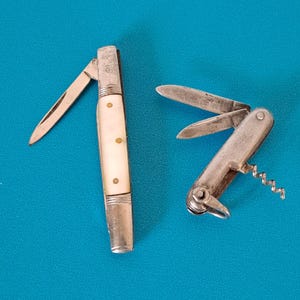 Peut inclure: Deux couteaux de poche pliants. Le couteau de gauche présente un manche de couleur crème avec trois goupilles en laiton et une lame déployée. Le couteau de droite est entièrement métallique, avec deux lames déployées, un tire-bouchon et un anneau.