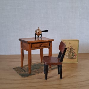 Puede incluir: Juego de muebles de madera en miniatura que incluye una mesa pequeña con un cajón, una silla y una alfombra decorativa. Una pequeña olla de color cobre se encuentra sobre la mesa. Una pequeña caja con una imagen de personas está al lado de la silla.