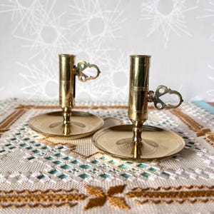 Peut inclure: Deux bougeoirs anciens en laiton. Chacun a une base ronde, une tige fine et un sommet cylindrique pour une bougie. Une poignée décorative est fixée sur le côté. Les bougeoirs sont sur une surface à motifs.