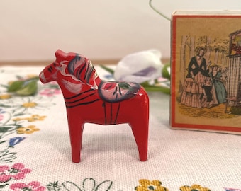 May include: A small, hand-painted wooden Dala horse in bright red with decorative floral and stripe details. The horse has a white, gray, and black mane and saddle. A vintage matchbox with a detailed illustration is in the background.