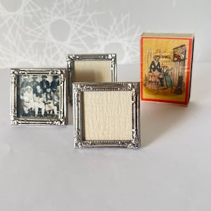 Może przedstawiać: Miniaturowe srebrne ramki do zdjęć z ozdobnymi detalami. Jedna z ramek zawiera czarno-białe zdjęcie grupy osób. Obecne jest również zabytkowe pudełko zapałek z kolorową ilustracją rodziny.