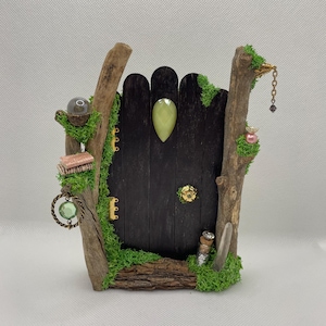May include: A miniature fairy door made of dark wood, adorned with moss, twigs, and decorative elements. The door features a green gemstone, a small bottle, and a hanging chain. A small book and a crystal ball are also visible.