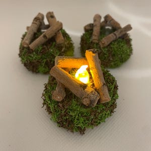 May include: Three miniature fairy garden campfires. Each campfire features a small, round base of green moss, topped with small wooden logs arranged to resemble a campfire. One campfire has a small, glowing LED light to simulate a fire.