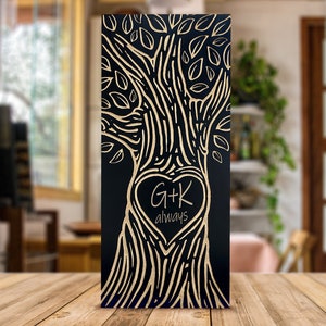 Op de afbeelding: Een zwart houten wandkunstwerk met een boomontwerp en goudkleurige accenten. De boom heeft een hartvormig centrum met de initialen "G+K" en het woord "always". Het kunstwerk wordt op een houten tafel getoond.