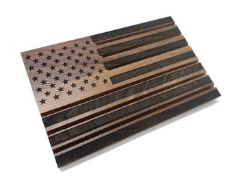 May include: Wooden American flag with a distressed finish. The flag features engraved stars in the upper left corner and alternating dark and light wood stripes. The flag is displayed on a white background.