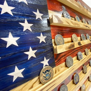 May include: Wooden American flag with red and blue painted stripes and white stars. The flag has several shelves holding various challenge coins. The wood has a distressed finish.