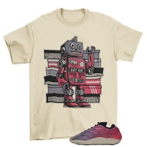 Pu&ograve; includere: Una maglietta beige con un robot dei cartoni animati che indossa una chiave inglese e tiene un cartello che dice "GOT EM". Il robot &egrave; rosso, rosa e blu. Sono anche raffigurate un paio di scarpe da ginnastica rosa e grigie.