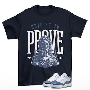 May include: A navy blue t-shirt with the text "NOTHING TO PROVE" in white lettering. A graphic of a skull wearing a crown and holding scales is centered on the shirt. A pair of white and blue sneakers are shown below the shirt.
