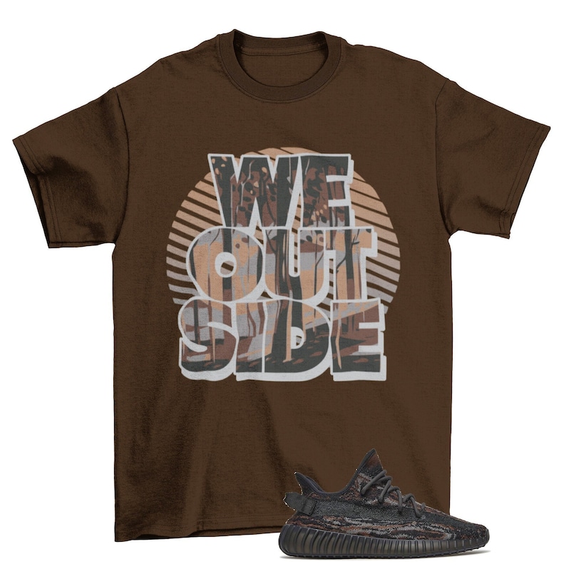 May include: A brown t-shirt with the text "WE OUTSIDE" printed on it in a white and gray font. The text is surrounded by a brown and gray graphic of trees. A pair of black and brown sneakers are shown below the t-shirt.