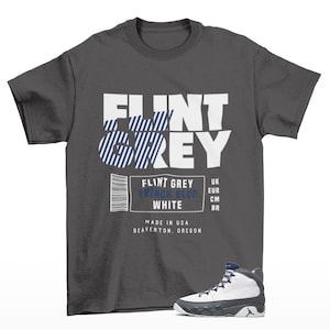 May include: A dark gray t-shirt with the words "FLINT GREY" in white block letters. The word "GREY" has blue and white striped accents. Below the text is a label with color names and a barcode. A white, gray, and blue sneaker is in the lower right.