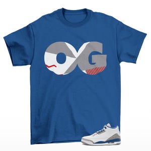 May include: A royal blue short-sleeve t-shirt featuring a large graphic of the letters OG in grey, white, and red with a geometric pattern. Below the shirt, a white athletic sneaker with grey elephant print accents and blue details is visible.
