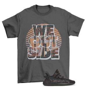 May include: A dark gray t-shirt with the text "WE OUTSIDE" printed in white with a brown and orange forest scene behind the text. A pair of black and brown sneakers with a camouflage pattern are shown below the t-shirt.