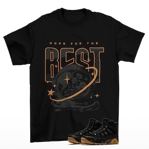 Puede incluir: Camiseta negra con el texto "HOPE FOR THE BEST" sobre un gráfico de un casco de astronauta con flores. Debajo, zapatillas negras con suelas marrones.