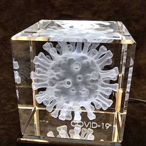May include: A clear glass cube with a 3D etched image of a coronavirus inside. The cube is on a black base and has the text "COVID-19" etched on the bottom.