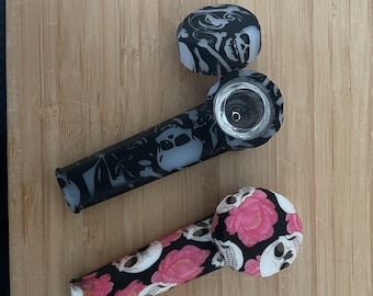May include: Two silicone smoking pipes with skull and floral designs. One pipe is black with white skulls and the other is black with pink flowers and white skulls.