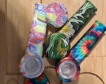 May include: Four colorful silicone smoking pipes with different patterns, including tie-dye, geometric, and floral designs. The pipes have clear glass bowls and are arranged on a wooden surface.
