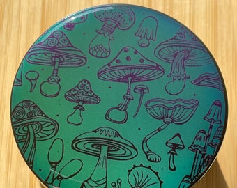 Puede incluir: Un recipiente redondo de metal plateado con un acabado iridiscente verde y morado. El recipiente está decorado con un dibujo de línea en blanco y negro de varios hongos.