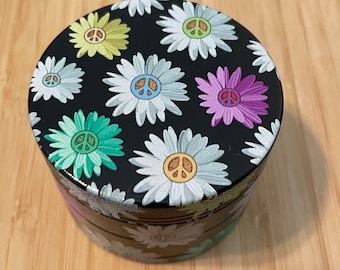 Può includere: Un contenitore rotondo e nero con un coperchio decorato con margherite bianche, ognuna con un simbolo di pace in vari colori. Il contenitore è posto su una superficie di legno marrone chiaro.