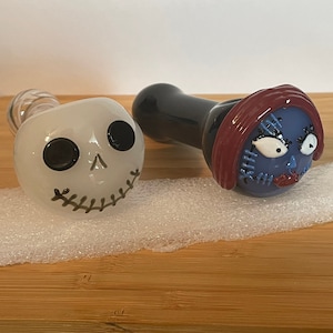 May include: Two glass pipes with character faces. One pipe is white with a black skull and stitched mouth. The other pipe is black with a blue face with red hair and stitched details.