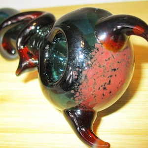 May include: A hand-blown glass pipe with a swirling, spiral design. The pipe features dark green, black, and red colours. The bowl has a speckled red pattern. The mouthpiece and end are curved.