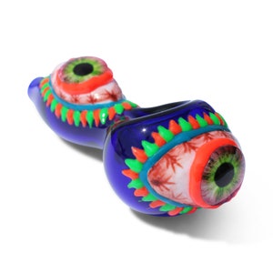 May include: A blue glass pipe with a unique design. The pipe features two large, detailed eye designs with green irises, red and orange accents, and a cannabis leaf pattern. The pipe is approximately 5 inches long.
