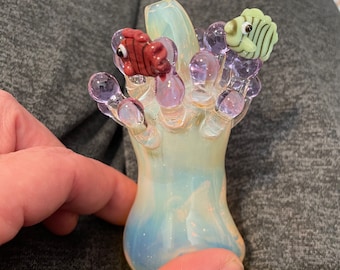 May include: A glass figurine of a coral reef with two small fish, one red and one green, on top. The coral is a light purple color and the base is a translucent white with a blue and white swirl.