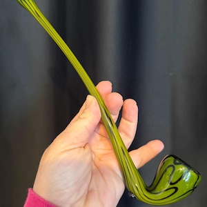 Puede incluir: Una pipa de vidrio verde con diseños negros en espiral. El largo tallo tiene rayas negras verticales. El cuenco es redondeado con una forma curva. La pipa se sostiene en una mano sobre un fondo oscuro.