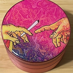 Puede incluir: Un molinillo de metal rosa con un dise&ntilde;o psicod&eacute;lico colorido que presenta dos manos que se alcanzan, una de las cuales sostiene un porro. El dise&ntilde;o recuerda a La Creaci&oacute;n de Ad&aacute;n de Miguel &Aacute;ngel.