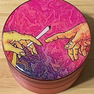 Puede incluir: Un molinillo rosa y morado con un dise&ntilde;o psicod&eacute;lico que presenta dos manos que se alcanzan, una de las cuales sostiene un cigarrillo blanco. El molinillo tiene una tapa de metal y una base texturizada.