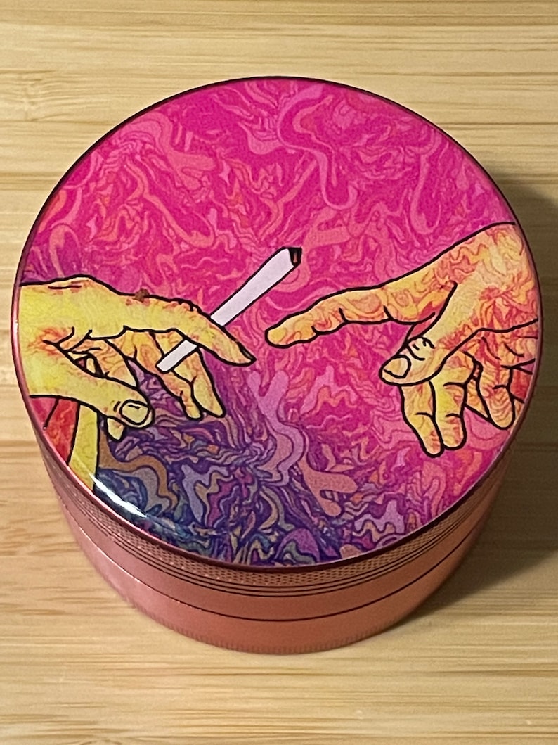Puede incluir: Un molinillo rosa y morado con un dise&ntilde;o psicod&eacute;lico que presenta dos manos que se alcanzan, una de las cuales sostiene un cigarrillo blanco. El molinillo tiene una tapa de metal y una base texturizada.