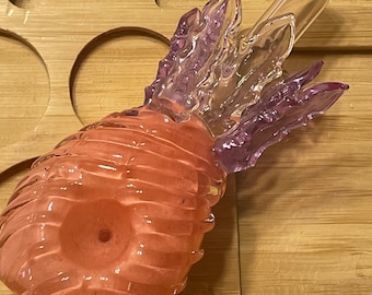 May include: A pink and clear glass pineapple shaped pipe with a removable mouthpiece. The pipe has a textured surface and a detailed pineapple design.