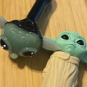 May include: Two glass pipes, one shaped like a green alien character with black eyes and a beige robe, and the other shaped like a green alien head with black eyes and a black stem.