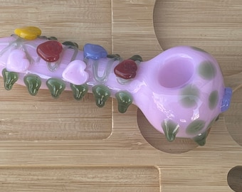 May include: A pink glass pipe with green and white accents. The pipe has a bowl on one end and a mouthpiece on the other. The pipe is decorated with colorful hearts and flowers.