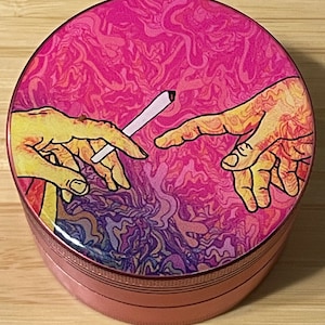 Puede incluir: Un molinillo rosa y morado con un dise&ntilde;o psicod&eacute;lico que presenta dos manos que se alcanzan, una de ellas sosteniendo un porro. El dise&ntilde;o recuerda a La Creaci&oacute;n de Ad&aacute;n de Miguel &Aacute;ngel.