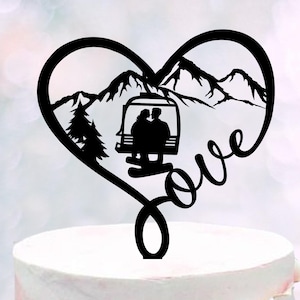 May include: Black heart-shaped cake topper featuring a mountain range, a ski lift with a couple, and the word "love". A pine tree is on the left side. The topper is placed on a white cake with a pink layer.