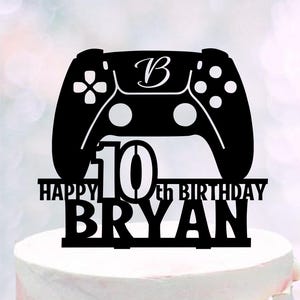 May include: Black cake topper in the shape of a video game controller. The topper reads "HAPPY 10th BIRTHDAY BRYAN" with the letter "B" and the number "10" in the design. The topper is placed on top of a white cake.