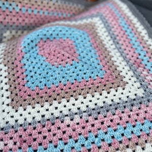 Pode incluir: Uma manta de crochê com um padrão geométrico em tons de rosa, azul, cinza e branco.