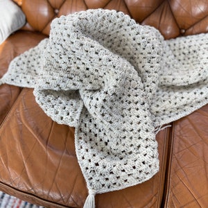 May include: A white crocheted throw blanket with a fringe detail, draped over a brown leather couch.
