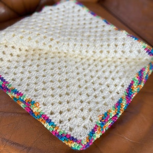 Pode incluir: Uma manta de crochê branca com uma borda colorida. A borda é feita de fios de diferentes cores, incluindo rosa, laranja, amarelo, verde, azul e roxo.