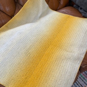 Pode incluir: Uma manta de crochê com um degradê de branco para amarelo.