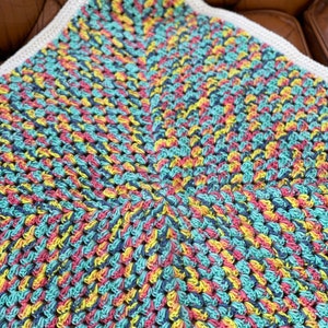 Pode incluir: Uma manta de crochê colorida com uma borda branca. A manta é feita com uma variedade de cores, incluindo rosa, azul, amarelo e verde.