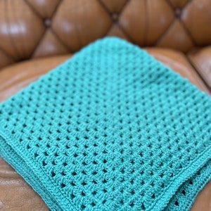 Pode incluir: Uma manta de crochê turquesa com um padrão de ponto quadrado. A manta está dobrada e colocada em um sofá de couro marrom.