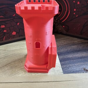 May include: A 3D-printed red castle tower with a crenelated top and a small rectangular window. The tower has a cylindrical body with vertical lines and a rectangular base. The object is made of plastic.