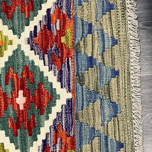May include: Close-up of a colorful rug with a geometric pattern. The rug has a fringe border and is made of wool. The colors in the rug are red, blue, green, and white.