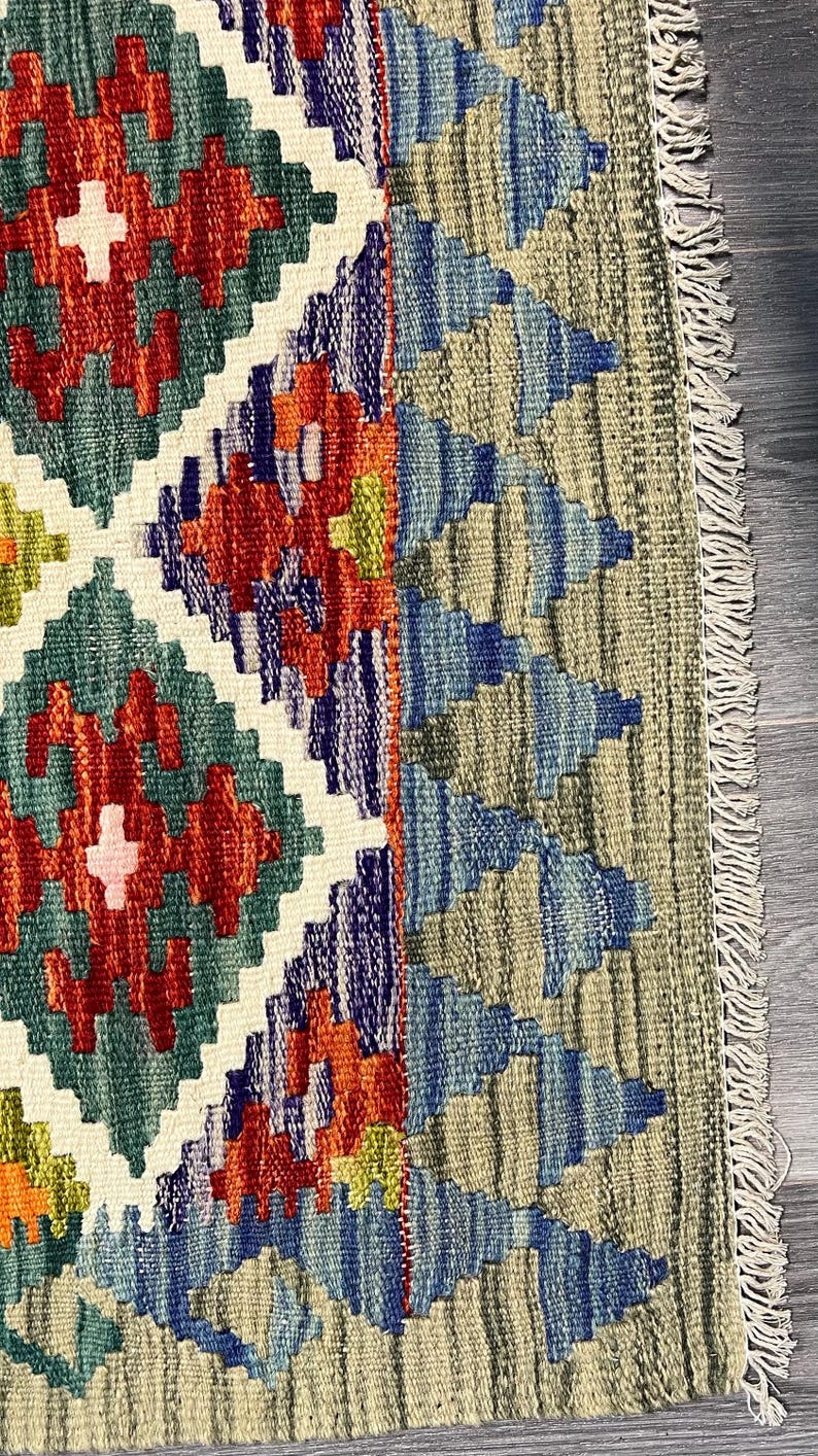 May include: Close-up of a colorful rug with a geometric pattern. The rug has a fringe border and is made of wool. The colors in the rug are red, blue, green, and white.