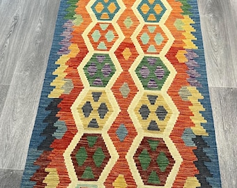 Könnte beinhalten: Ein rechteckiger, handgewebter Teppich mit geometrischem Muster. Der Teppich hat einen blauen Rand und ein zentrales Design aus sich wiederholenden sechseckigen Formen in Orange, Gelb, Grün und Lila. Der Teppich ist etwa 244 cm lang.