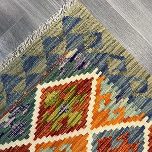 May include: A colorful rug with a geometric pattern in shades of blue, green, orange, and brown. The rug has a fringe border.