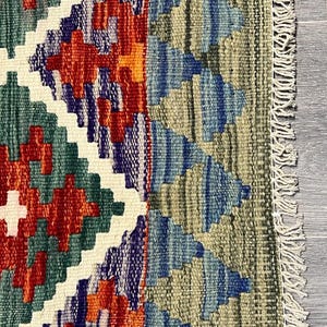 May include: A colorful rug with a geometric pattern in shades of red, blue, green, and white. The rug has a fringe border.